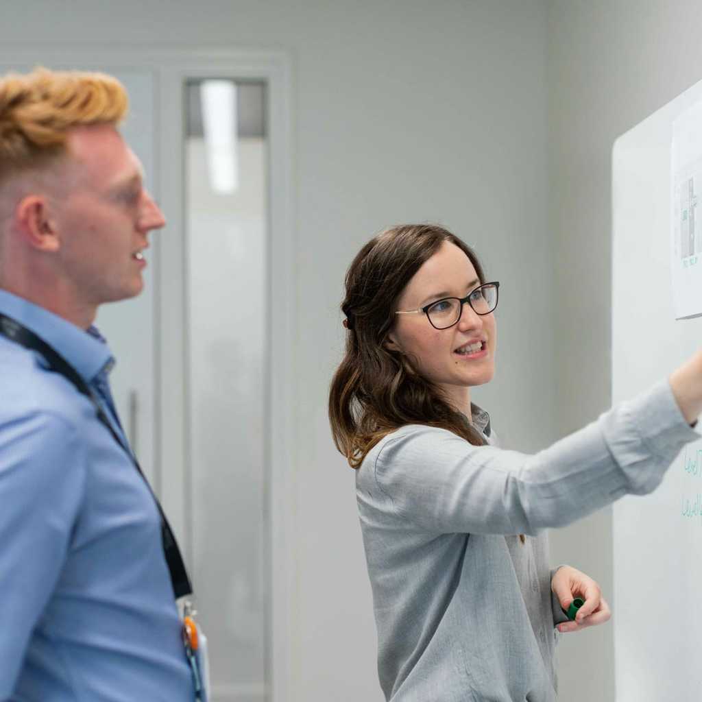 Två personer som står vid en white board. Den ena personen håller i en penna och visar något på tavlan till den andra.