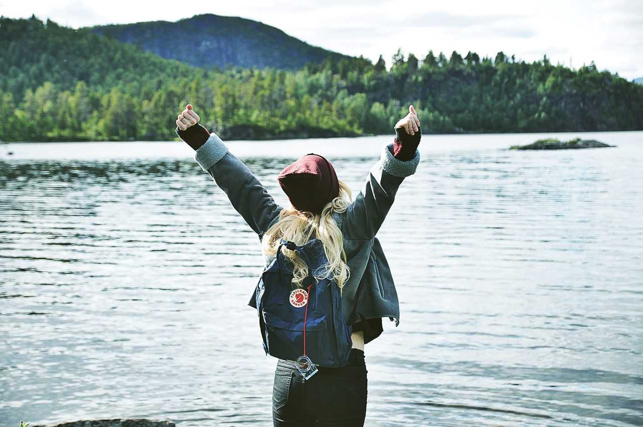Kvinnor ser ut över vatten med händerna i luften. Har röd mössa och blå kåken ryggsäck.