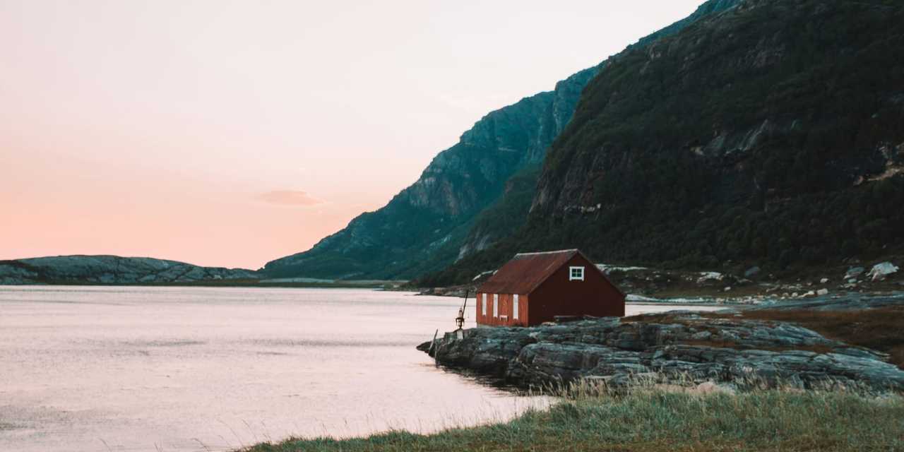 En röd stuga vid vattnet med ett berg i bakgrunden i solnedgången. 