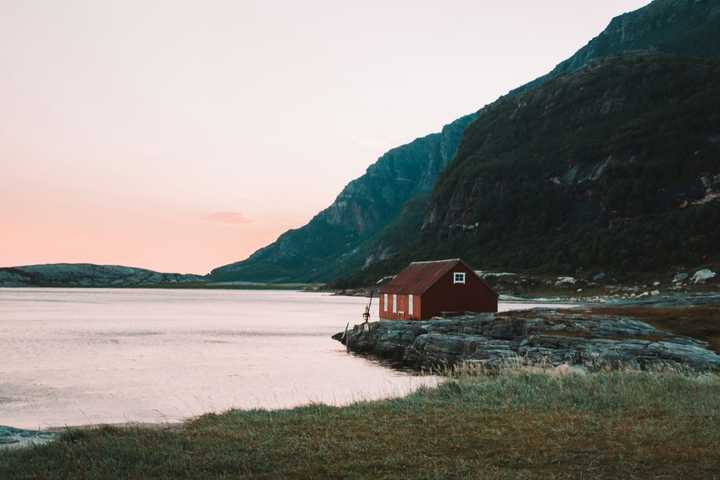 En röd stuga vid vattnet med ett berg i bakgrunden i solnedgången. 